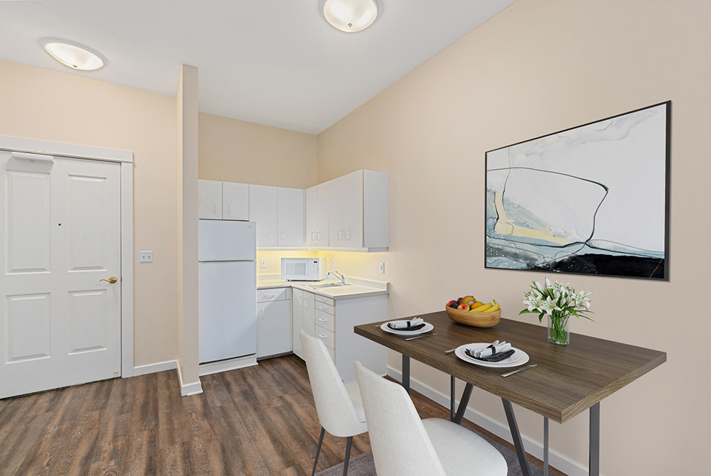 A small, modern kitchen and dining area in a 143 Conversion Apartment with white cabinets, a refrigerator, microwave, and light wood flooring. A dining table with two white chairs is set with plates and a fruit bowl, over which a minimalist painting hangs on the beige wall.