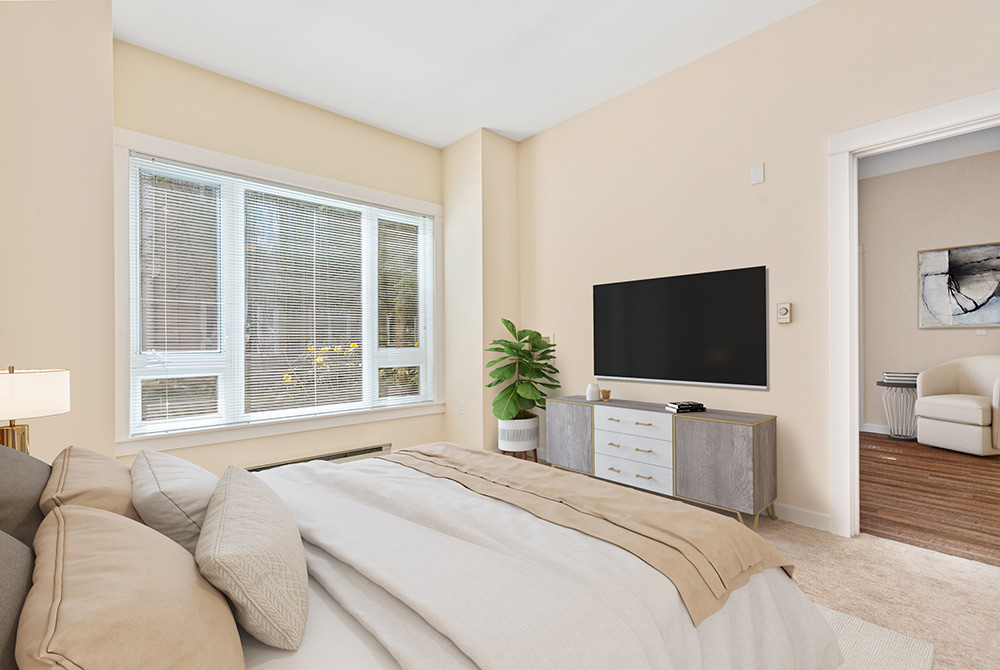 A bright and cozy bedroom in Conversion Apartment 143 features a large bed with beige bedding, a flat-screen TV on a light-colored dresser, and a potted plant beside it. Natural light pours in through the large window with blinds, while an open doorway leads to another room.