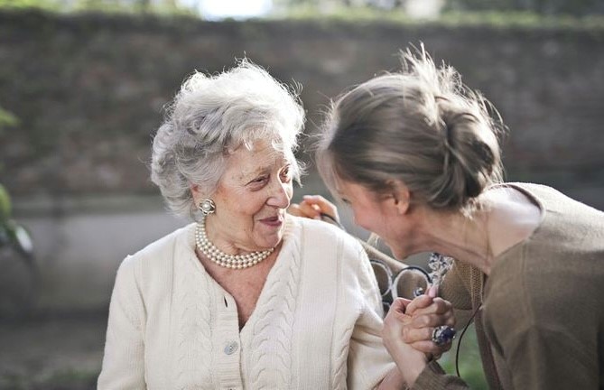 A smiling elderly woman with short, curly gray hair and wearing a white cardigan and pearl necklace is holding hands and sharing a joyful moment with a younger woman with light brown hair in a casual outfit. They are outdoors, embracing the highs amidst dementia-related lows in the blurred background.